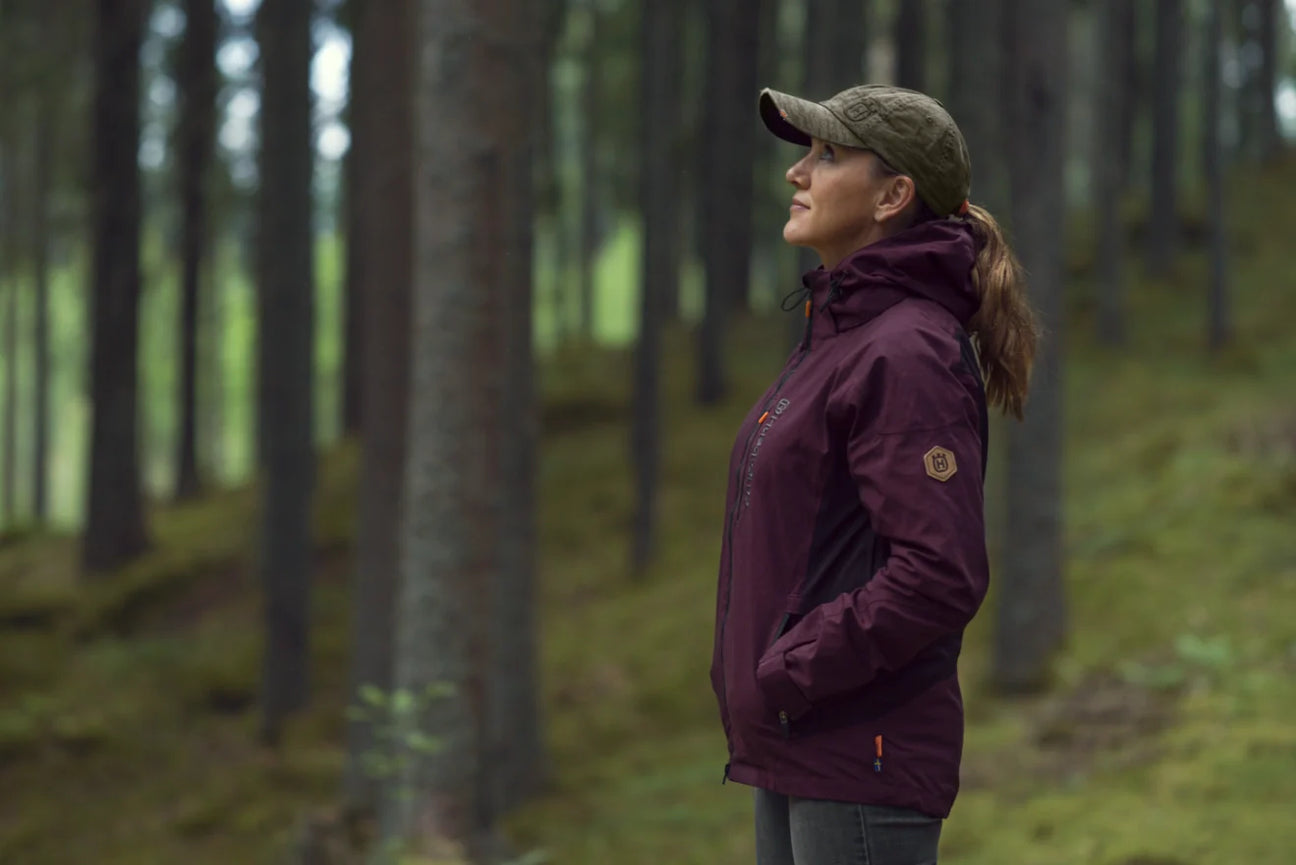 Husqvarna Xplorer vaatteet: Nainen seisoo metsässä päällään tyylikäs ja käytännöllinen Husqvarna Xplorer kuoritakki, täydellinen valinta ulkoiluun ja seikkailuihin.
